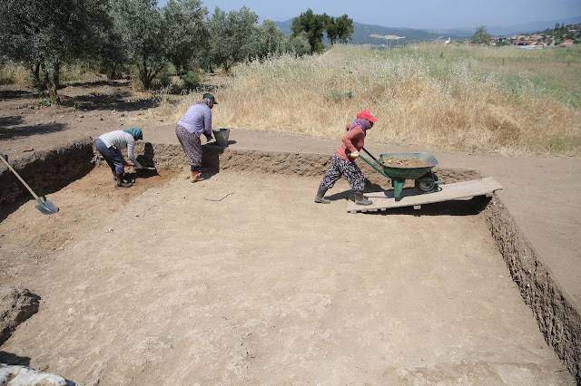 Αρχαιολόγοι εργάζονται σε ανασκαφικό τομέα στη Μαγνησία, αφαιρώντας στρώματα χώματος σε ανοιχτό, αγροτικό τοπίο.