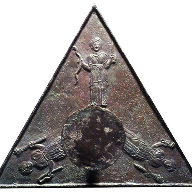 Τρίγωνο χάλκινο έλασμα με αιχμητές ακμές· το κάθε πλευρό σχηματίζει ισόπλευρο τρίγωνο. Στο κέντρο του τριγώνου, χαραγμένος ελαφρά ανάγλυφος, υπάρχει ένας κυκλικός δίσκος (ασπίδα ή κύκλος) που καταλαμβάνει το περισσότερο χώρο στο βάθος. Πάνω από τον κύκλο, σε προεξοχή προς τα πάνω, στέκεται όρθια μια γυναικεία μορφή: φορά μακριά, πλούσια αναδιπλωμένη χιτώνα που σκεπάζει το σώμα της από τη μέση έως τις αστράγαλους. Έχει ζωσμένη τη μέση και στο κεφάλι φέρει κάποιο είδος στεφάνου. Το αριστερό χέρι της είναι λυγισμένο προς τα πάνω, όπου απ’ τα δάχτυλά της κρέμεται ένα φιδίσιο αντικείμενο (πιθανότατα όφις ή φίδι), ενώ το δεξί της χέρι, επίσης λυγισμένο, κρατάει ίσως ένα ρόπαλο ή τυπική ραβδία. Το βλέμμα της στρέφεται ευθεία προς τα εμπρός.
Στη βάση του κεντρικού κύκλου, αριστερά και δεξιά, υπάρχουν δύο οριζόντια τοποθετημένες γυναικείες μορφές σχεδόν καθιστές και γερμένες λίγο προς τα έξω. Η αριστερή μορφή γέρνει προς τα αριστερά, φορά κι αυτή μακριά χιτώνα με πλούσιες πτυχώσεις και δένει με ζώνη τη μέση της. Στο δεξί της χέρι κρατάει λεπτό αντικείμενο (πιθανότατα κύλινδρο με επιγραφή ή ραβδί), ενώ το άλλο της χέρι ακουμπά χαλαρά στο έδαφος. Το κεφάλι της γέρνει ελαφρώς προς τα κάτω.
Η δεξιά μορφή καθισμένη γέρνει προς τα δεξιά· φορά επίσης μακριά, κοντά σε στυλ χιτώνα, με εμφανείς πτυχώσεις και ζώνη στη μέση. Στο αριστερό χέρι φαίνεται να κρατά κάποιο ρόπαλο ή μικρό δόρυ, ενώ το δεξί χέρι κρέμεται χαλαρά δίπλα στο σώμα της. Το κεφάλι της είναι στραμμένο ελαφρώς προς τα πίσω, δίνοντας την αίσθηση ότι κοιτά προς άλλη κατεύθυνση.
Όλα τα σώματα και τα πρόσωπα των θεοτήτων είναι σκαλισμένα με απαλές γραμμές και καθαρές λεπτομέρειες στις πτυχώσεις των ενδυμάτων, ενώ ο μεταλλικός δίσκος στο κέντρο παρουσιάζει φθορές και πατίνα από την πάροδο του χρόνου. Στο φόντο φαίνονται φθαρμένα επιγραφικά ή διακοσμητικά μοτίβα που διακοσμούν ολόγυρα το τρίγωνο. Η συνολική υφή είναι τραχιά, με μικρές εσοχές και ίχνη ξυσίματος, ενδεικτικά του αρχαίου καλλιτεχνικού χειροποίητου χαρακτήρα αυτού του μαγικού πλακιδίου.