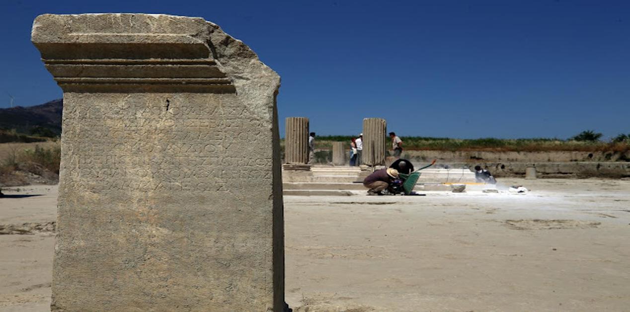Αρχαία λίθινη επιγραφή στη μαρμάρινη αγορά της Μαγνησίας, με φόντο αρχαιολόγους που συνεχίζουν τις ανασκαφές ανάμεσα σε κίονες.
