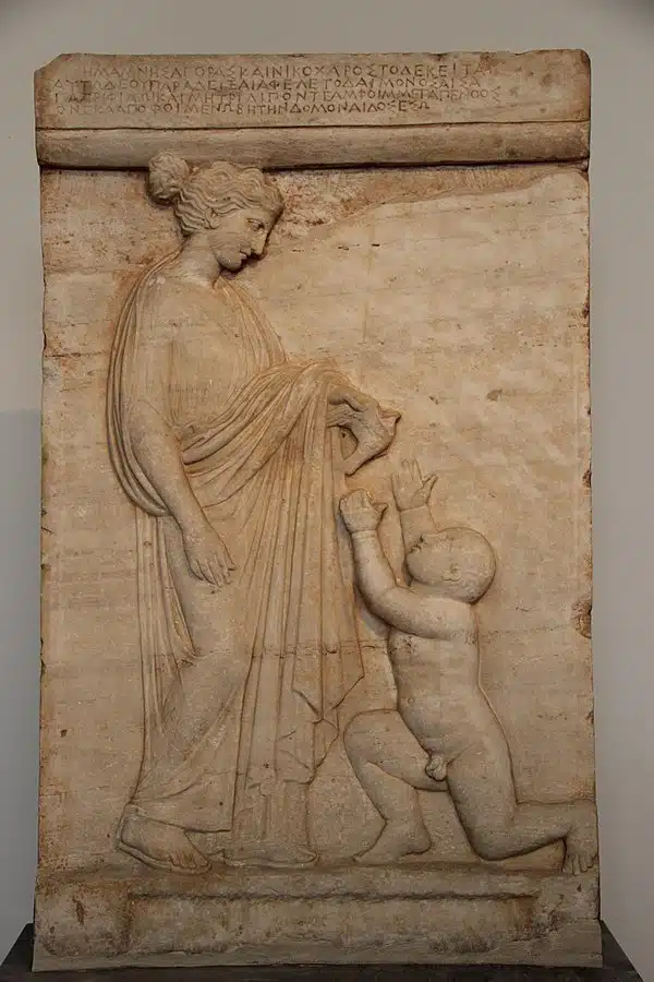 Ανάγλυφη αρχαία ελληνική ταφική στήλη. Εικονίζεται μια γυναίκα, όρθια με ιμάτιο, που κοιτάζει στοργικά ένα μικρό παιδί. Το παιδί, γυμνό και γονατιστό, υψώνει τα χέρια του προς τη γυναίκα και αγγίζει το ρούχο της, σε κίνηση αποχαιρετισμού. Στην κορυφή της στήλης διακρίνεται επιγραφή με αρχαία ελληνικά γράμματα. Το έργο αποπνέει τρυφερότητα, θλίψη και μητρική αγάπη.