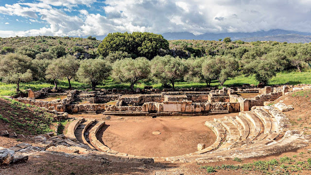 «Πανοραμική άποψη του αρχαίου θεάτρου της Άπτερας, με το ημικυκλικό κοίλο και την ορχήστρα να εντάσσονται αρμονικά στο μεσογειακό τοπίο με ελαιώνες και Λευκά Όρη στο βάθος.»