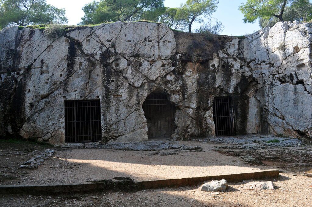 Εξωτερική όψη της Φυλακής του Σωκράτη στην Αθήνα, ένα λαξευμένο σπήλαιο με τρεις είσοδοι, οι δύο με σιδερένιες μπάρες.