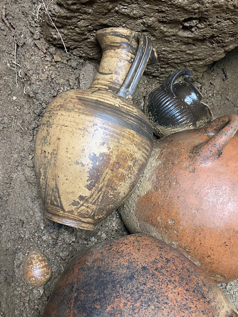 Κοντινή λήψη από ετρουσκικά αγγεία σε ταφικό θάλαμο στο San Giuliano, με ένα διακοσμημένο πιθάρι και ένα μαύρο μικρό αγγείο ανάμεσα στα κτερίσματα.