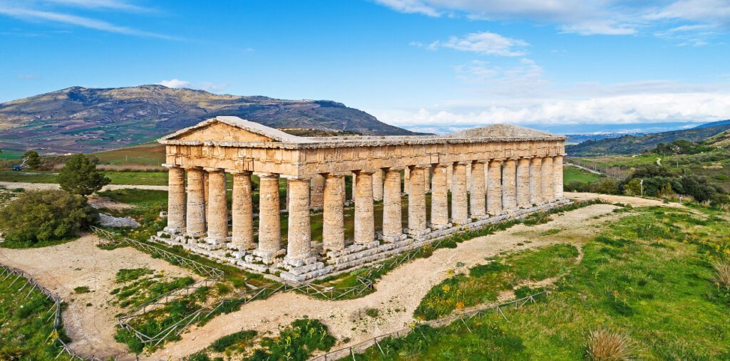 Η Ελληνική Αποίκιση στη Μεγάλη Ελλάδα - Ευρυγώνια άποψη του μεγάλου, ημιτελούς δωρικού ναού της Σέγεστας. Ο ναός, χωρίς οροφή, στέκεται σε έναν καταπράσινο λόφο με φόντο την ορεινή ύπαιθρο της Σικελίας κάτω από έναν γαλάζιο ουρανό.