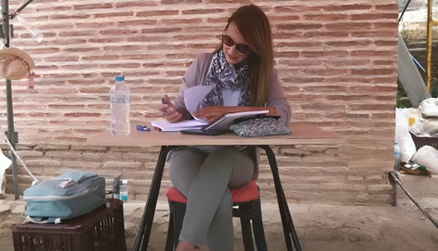 Γυναίκα αρχαιολόγος κάθεται σε γραφείο σε υπαίθριο χώρο ανασκαφής και καταγράφει δεδομένα στο ημερολόγιό της.