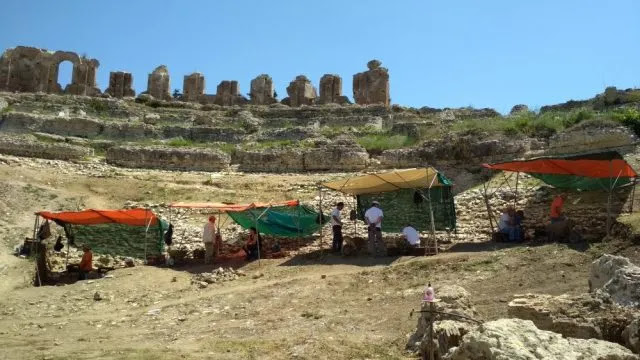 Αρχαιολόγοι εργάζονται σε ανασκαφή με προσωρινά σκίαστρα, μπροστά από ερείπια αρχαίου θεάτρου ή οχυρωματικού τείχους, κάτω από καθαρό ουρανό.