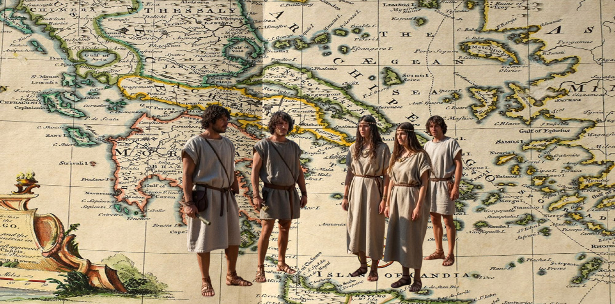 Η Γένεση των Ελλήνων - Αρχαία Ελληνικά Φύλα. Μια ομάδα πέντε ατόμων, ντυμένοι με παραδοσιακές αρχαιοελληνικές ενδυμασίες, απεικονίζονται μπροστά σε έναν ιστορικό χάρτη της Ελλάδας και του Αιγαίου. Ο χάρτης δείχνει γεωγραφικές περιοχές όπως η Θεσσαλία, η Μακεδονία και η Πελοπόννησος. Τα άτομα φορούν χιτώνες και σανδάλια, και μερικά έχουν αξεσουάρ όπως ζώνες και καλύμματα κεφαλής.
