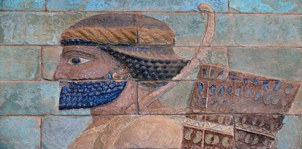 Οι Δέκα Χιλιάδες Αθάνατοι: Ο Θρύλος και η Πραγματικότητα της Στρατιάς του Ξέρξη - Μια λεπτομερής κοντινή λήψη ενός τμήματος της Ζωφόρου των Τοξοτών από το Παλάτι του Δαρείου Α' στα Σούσα, που απεικονίζει έναν Πέρση αθάνατο. Η φιγούρα εμφανίζεται σε προφίλ, στραμμένη αριστερά, με ένα εμφανές πλεγμένο γενειάδα σε μπλε χρώμα και σκούρα μαλλιά, καθώς και μια πλεγμένη κορδέλα στο κεφάλι. Φοράει έναν περίτεχνο, σχεδιασμένο χιτώνα και αυτό που φαίνεται να είναι μια φαρέτρα στην πλάτη του με ένα εμφανές ανακλαστικό τόξο. Το έργο τέχνης αποτελείται από πολύχρωμα εφυαλωμένα τούβλα, δείχνοντας την περίπλοκη λεπτομέρεια και διατήρηση της αρχαίας περσικής τέχνης.