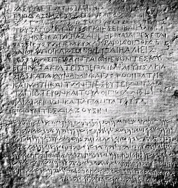Κοντινή ασπρόμαυρη φωτογραφία μιας αρχαίας πέτρινης επιγραφής. Το πάνω μέρος της πέτρας έχει κείμενο χαραγμένο σε αρχαία Ελληνικά κεφαλαία γράμματα, ενώ το κάτω μέρος έχει κείμενο σε Αραμαϊκή γραφή. Η πέτρα είναι φθαρμένη από τον χρόνο, αλλά τα γράμματα είναι ευδιάκριτα.