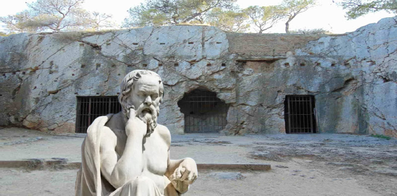 Φυλακή του Σωκράτη: Ιστορία, αρχαιολογικά ευρήματα και άγνωστες πτυχές - Άγαλμα του Σωκράτη μπροστά από τη Φυλακή του Σωκράτη στην Αθήνα, ένα σπήλαιο με τρεις εισόδους, δύο εκ των οποίων έχουν μεταλλικές μπάρες.