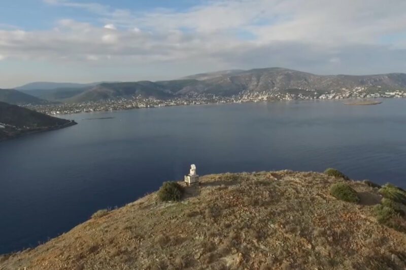 Ο Κολοσσός του Πόρτο Ράφτη σε ξηρό, καφέ λόφο με θάμνους, με φόντο έναν απέραντο γαλάζιο κόλπο και οικισμούς απέναντι, κάτω από συννεφιασμένο ουρανό.