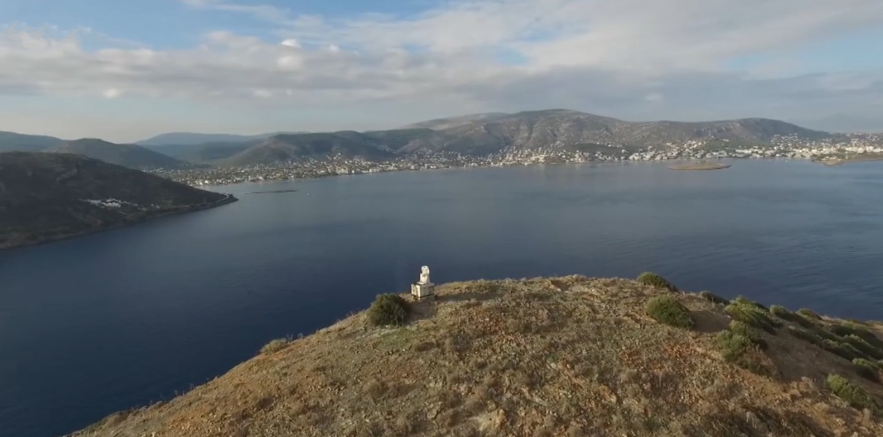 Ο Κολοσσός του Πόρτο Ράφτη σε ξηρό, καφέ λόφο με θάμνους, με φόντο έναν απέραντο γαλάζιο κόλπο και οικισμούς απέναντι, κάτω από συννεφιασμένο ουρανό.