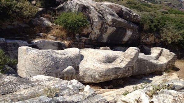 Ο Κούρος του Απόλλωνα: Αρχαίο Μυστήριο και Ιστορική Κληρονομιά της Νάξου - Ένα μεγάλο, οριζόντιο, ημιτελές άγαλμα Κούρου από λευκό μάρμαρο βρίσκεται στο έδαφος ενός βραχώδους λατομείου. Το άγαλμα, το οποίο φαίνεται να είναι ημιτελές, απεικονίζει μια ανθρώπινη μορφή σε ύπτια θέση. Γύρω του υπάρχουν μεγάλοι, ακανόνιστοι βράχοι και μερικοί πράσινοι θάμνοι, ενώ ο ουρανός δεν είναι ορατός.