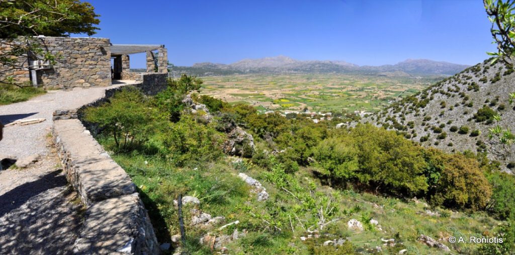 Δικταίο Άντρο: Ανακαλύψτε το Σπήλαιο του Δία στην Κρήτη - Μια γραφική πέτρινη διαδρομή οδηγεί προς την έξοδο του Δικταίου Άντρου, με θέα στο καταπράσινο Οροπέδιο Λασιθίου και τα γύρω βουνά της Κρήτης κάτω από έναν καθαρό, γαλανό ουρανό.
