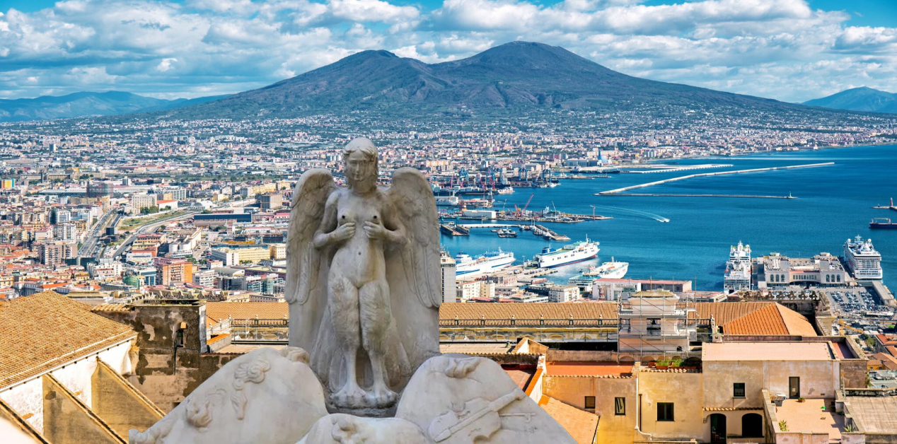 Παρθενόπη: Ο Μύθος της Αρχαίας Ελληνίδας Σειρήνας που Έγινε η Ψυχή της Νάπολης - Πανοραμική άποψη της πόλης της Νάπολης με τον Βεζούβιο στο βάθος. Σε πρώτο πλάνο, στις στέγες ενός κτιρίου, βρίσκεται ένα πέτρινο άγαλμα της Σειρήνας Παρθενόπης που κοιτάζει προς την πόλη.