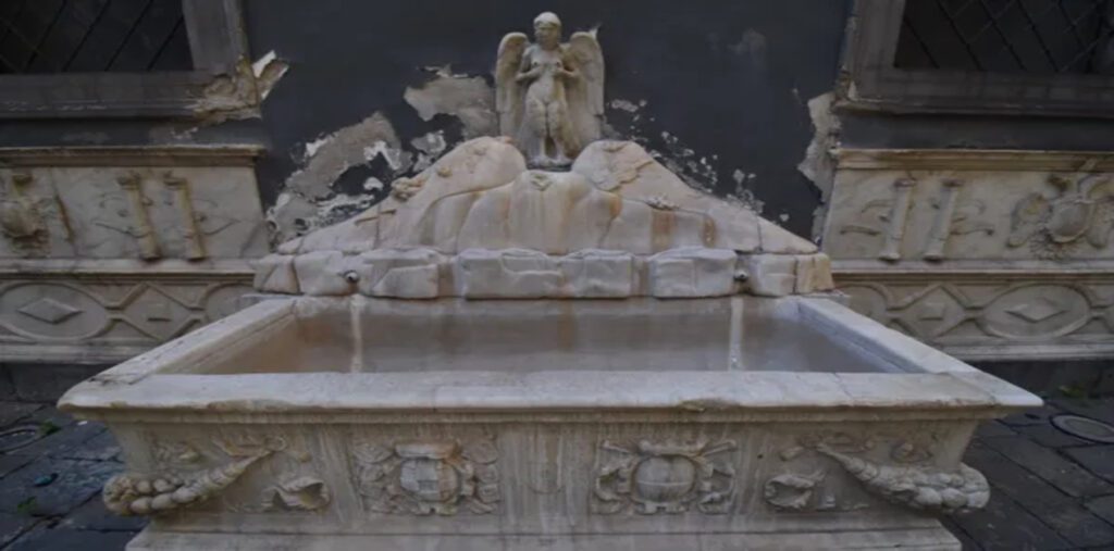 Παλιά μαρμάρινη κρήνη, η Fontana della Spinacorona, σε κοντινό πλάνο. Στο κέντρο, ένα γλυπτό αναπαριστά τη φτερωτή Σειρήνα Παρθενόπη πάνω από μια αναπαράσταση του Βεζούβιου.