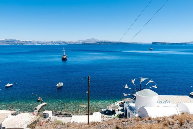 Πανοραμική θέα από τη Θηρασιά με παραδοσιακό ανεμόμυλο, λευκά κτίρια και βαθυγάλαζα νερά απέναντι από τη Σαντορίνη.