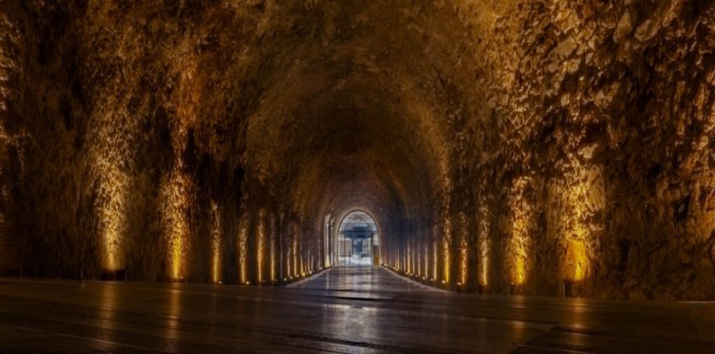 Εσωτερική άποψη ενός μακρύ, θολωτού πέτρινου τούνελ με ξύλινο πάτωμα, φωτισμένο από κάτω προς τα πάνω από κίτρινα φώτα που τονίζουν την υφή των τοίχων. Στο βάθος διακρίνεται μια φωτεινή έξοδος.