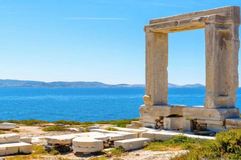 Πορτάρα Νάξου: Ταξίδι στο Χρόνο και την Ιστορία - Μνημείο Πορτάρα Νάξου, Ελλάδα. Η φωτογραφία απεικονίζει τη μεγάλη μαρμάρινη πύλη σε έναν βραχώδη λόφο, με φόντο τη γαλάζια θάλασσα και τα βουνά. Ο ουρανός είναι καθαρός και φωτεινός.