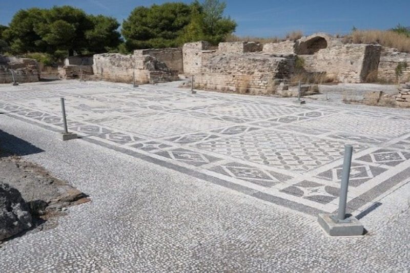 Η άγνωστη πισίνα του Νέρωνα στην Κόρινθο - Ψηφιδωτό δάπεδο αρχαίας Ρωμαϊκής πισίνας του Νέρωνα στην Κόρινθο, με μαύρα και άσπρα γεωμετρικά σχέδια και εικονιστικά πλαίσια, ανάμεσα σε πέτρινους τοίχους και πράσινο φόντο.