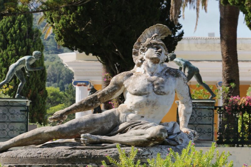 Η Αληθινή Σημασία Πίσω από το Όνομα «Αχιλλέας» - Μαρμάρινο άγαλμα του αρχαίου Έλληνα ήρωα Αχιλλέα, τη στιγμή του θανάσιμου τραυματισμού του. Είναι ξαπλωμένος σε έναν βράχο, φορώντας μόνο την περικεφαλαία του. Το άγαλμα βρίσκεται σε εξωτερικό χώρο, μέσα σε έναν ολάνθιστο κήπο με δέντρα και άλλα γλυπτά στο βάθος.