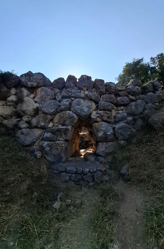 Η αρχαία μυκηναϊκή γέφυρα του Αρκαδικού, χτισμένη με μεγάλες, ακανόνιστες πέτρες (κυκλώπεια τοιχοποιία). Στο κέντρο της διακρίνεται η μικρή, τριγωνική δίοδος της γέφυρας, μέσα από την οποία φαίνεται το φως. Ένα μονοπάτι οδηγεί προς τη δίοδο, ενώ ο ήλιος φωτίζει τον ουρανό πάνω από την κορυφή της.