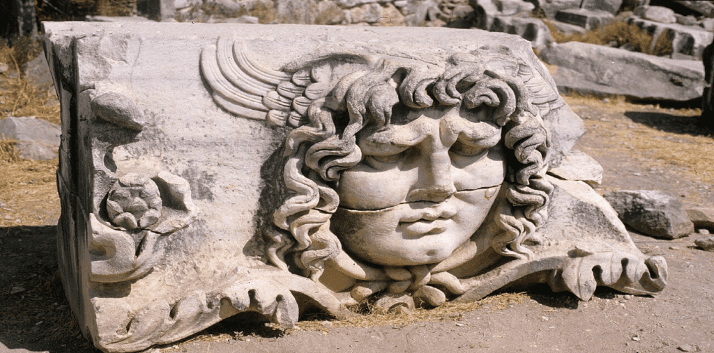 Μαρμάρινη κεφαλή Γοργόνας (Γοργόνειο) σε κοντινό πλάνο, πεσμένη στο έδαφος ανάμεσα σε αρχαία ερείπια. Το γλυπτό έχει σπασίματα και φθορές, αλλά διακρίνονται καθαρά τα κυματιστά μαλλιά, τα φτερά πάνω από το μέτωπο και τα έντονα χαρακτηριστικά του προσώπου.