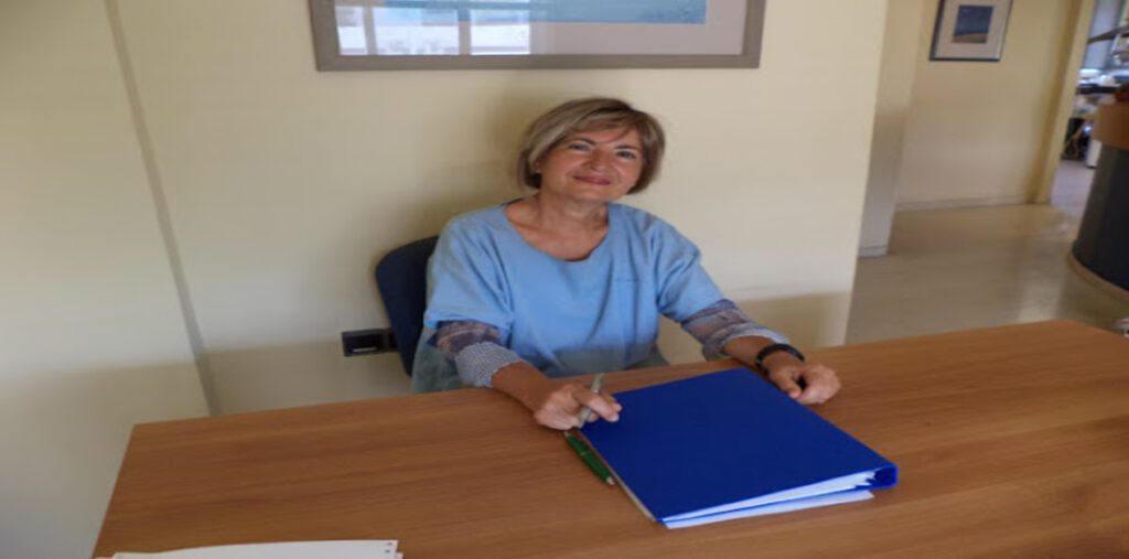 Φωτογραφία της Δήμητρας Τυλλιανάκη, χειρουργού οδοντιάτρου, καθώς κάθεται πίσω από ένα ξύλινο γραφείο με έναν μπλε φάκελο μπροστά της. Η κ. Τυλλιανάκη φοράει ένα γαλάζιο πουκάμισο, έχει κοντά, ξανθά μαλλιά και κοιτάει προς την κάμερα.