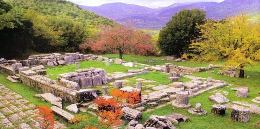 Τα πέτρινα ερείπια του αρχαιολογικού χώρου της Λυκόσουρας απλωμένα σε μια έκταση με πλούσια, πράσινη χλόη. Στο φόντο υψώνονται λόφοι και βουνά με πυκνή βλάστηση και δέντρα με φθινοπωρινά φύλλα σε κίτρινες και κόκκινες αποχρώσεις.