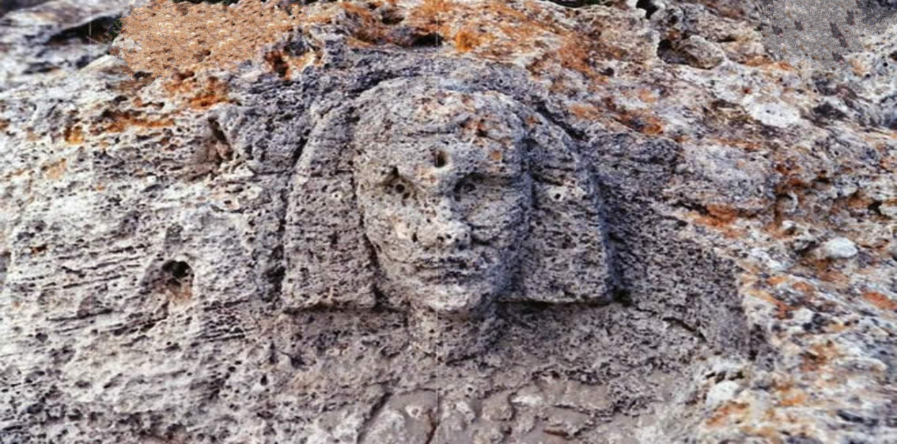 Η Πυραμίδα στα Βιγκλάφια Λακωνίας και η Μυστηριώδης Σφίγγα - Κοντινό πλάνο ενός προσώπου σκαλισμένου σε έναν πορώδη, διαβρωμένο βράχο. Το πρόσωπο έχει αρχαϊκά χαρακτηριστικά και ένα σχηματοποιημένο κάλυμμα κεφαλής ή μαλλιά που το πλαισιώνουν.