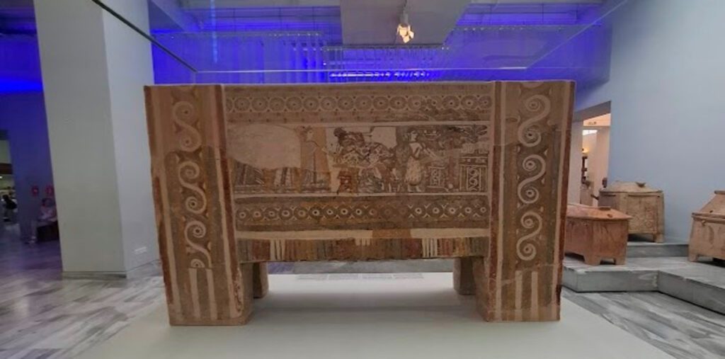 Η πίσω πλευρά της ζωγραφισμένης Σαρκοφάγου της Αγίας Τριάδας. Στο κέντρο, ένας ταύρος είναι δεμένος σε τραπέζι για θυσία, με μορφές τριγύρω. Αριστερά, τέσσερις γυναίκες σε πομπή πλησιάζουν ένα ιερό.