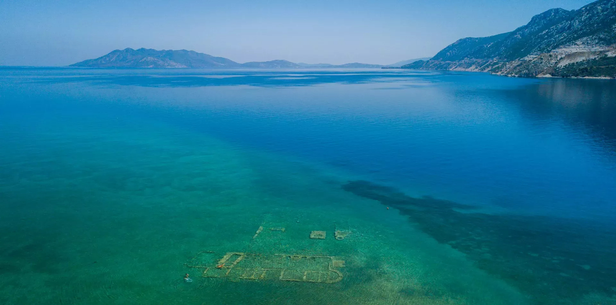 Βυθισμένη Πολιτεία Αρχαίας Επιδαύρου - Αεροφωτογραφία της Βυθισμένης Πολιτείας της Επιδαύρου. Στο προσκήνιο, τα θεμέλια αρχαίων κτιρίων είναι ορατά κάτω από τα διάφανα, γαλαζοπράσινα νερά. Στο βάθος, η θάλασσα βαθαίνει και διακρίνονται βουνά στον ορίζοντα.