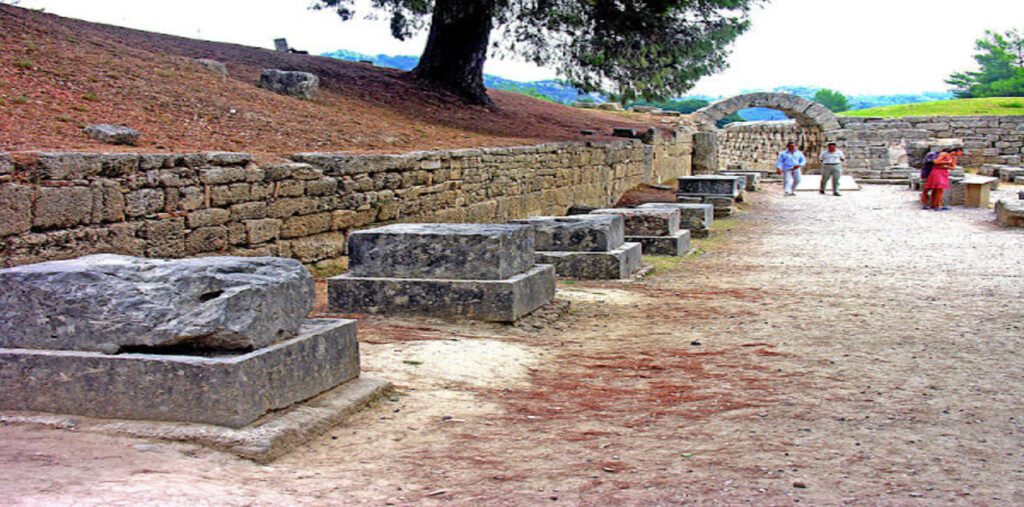 Οι πέτρινες βάσεις των αγαλμάτων των Ζανών σε σειρά, στον αρχαιολογικό χώρο της Αρχαίας Ολυμπίας, με την είσοδο του σταδίου στο βάθος.