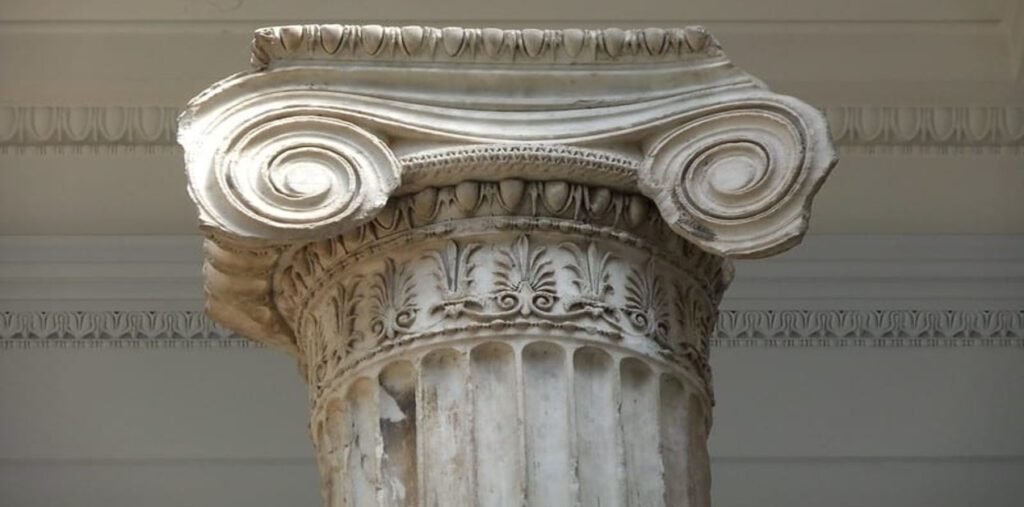 Φωτογραφία που δείχνει ένα κοντινό πλάνο ενός λευκού μαρμάρινου ιωνικού κιονόκρανου, το οποίο διαθέτει δύο μεγάλες, καμπυλωτές σπείρες και περίτεχνα ανάγλυφα φυτικά μοτίβα στη βάση του. Το κιονόκρανο είναι τοποθετημένο πάνω σε τμήμα ενός κίονα.