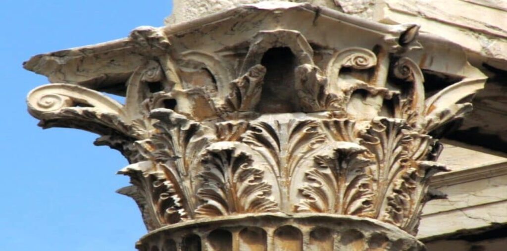 Μία φωτογραφία που δείχνει ένα κοντινό πλάνο ενός μαρμάρινου κορινθιακού κιονόκρανου. Το κιονόκρανο καλύπτεται από ανάγλυφα φύλλα ακάνθου και μικρές έλικες. Το μάρμαρο έχει πατίνα και φθορές από το χρόνο, με φόντο τον γαλάζιο ουρανό.