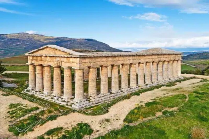 Πώς η αρχαία Ελληνική αρχιτεκτονική διαμόρφωσε τον σύγχρονο κόσμο - Πανοραμική άποψη ενός καλοδιατηρημένου αρχαίου Ελληνικού ναού Δωρικού ρυθμού. Ο ναός βρίσκεται σε έναν καταπράσινο λόφο στη Σικελία, με σκούρους πράσινους θάμνους και ένα μονοπάτι να οδηγεί προς αυτόν. Στο βάθος, λόφοι και βουνά φαίνονται κάτω από έναν γαλάζιο ουρανό με σύννεφα.