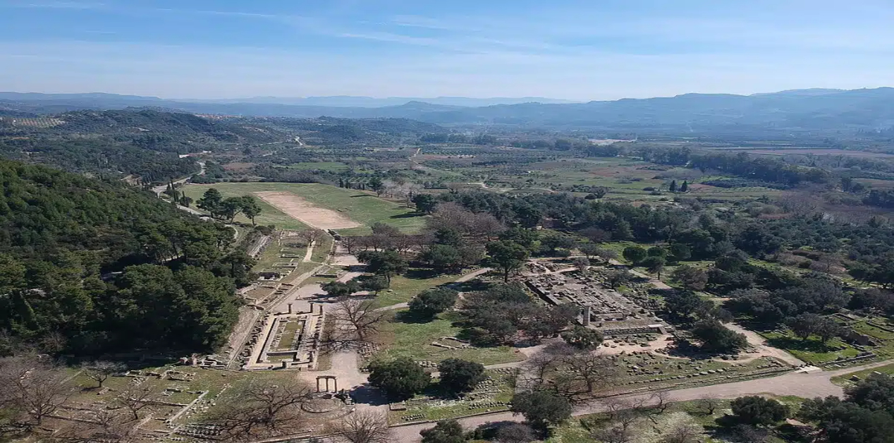 Η χαμένη λίμνη της Αρχαίας Ολυμπίας - Εναέρια όψη της Αρχαίας Ολυμπίας με ερείπια ναών και το αρχαίο στάδιο, περιτριγυρισμένα από πράσινο τοπίο και μακρινούς λόφους.