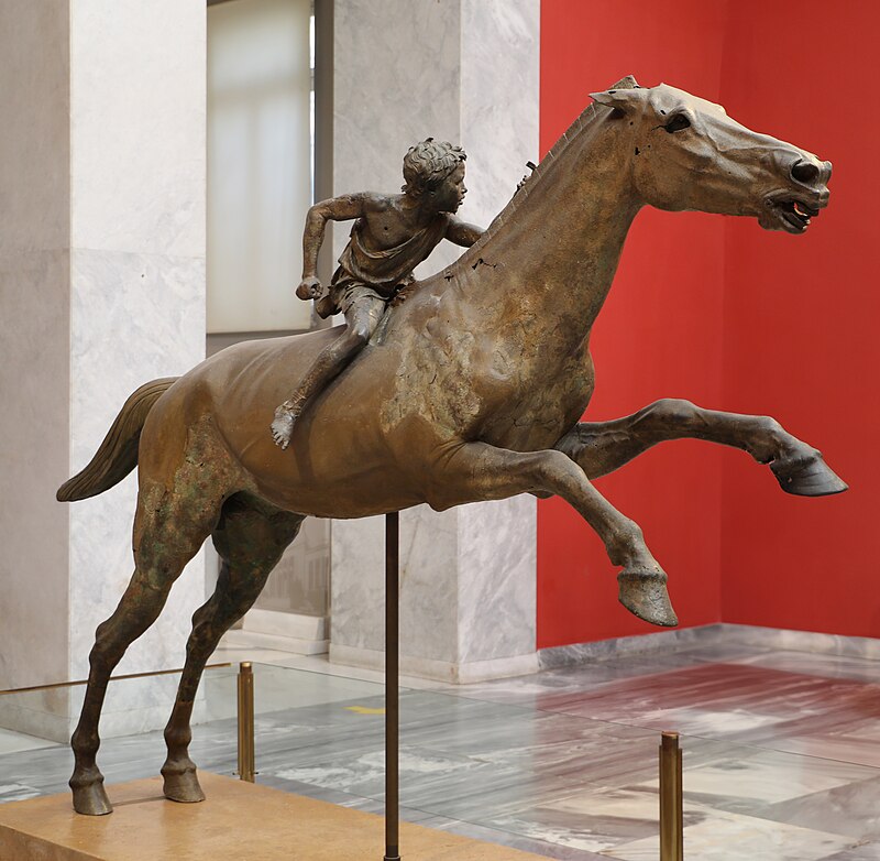 Πλάγια όψη του χάλκινου αγάλματος Ιππέας του Αρτεμισίου, που εκτίθεται σε μουσείο. Το άλογο είναι σε καλπασμό, με τον νεαρό αναβάτη να κοιτάζει πίσω. Το φόντο περιλαμβάνει κόκκινο τοίχο και μαρμάρινες κολώνες.