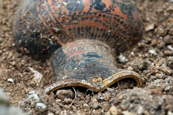 Κοντινή λήψη ενός αρχαιοελληνικού μελανόμορφου αγγείου (οινοχόη) με χρυσή επένδυση στο στόμιο, μισοθαμμένου στο χώμα κατά τη διάρκεια ανασκαφής.