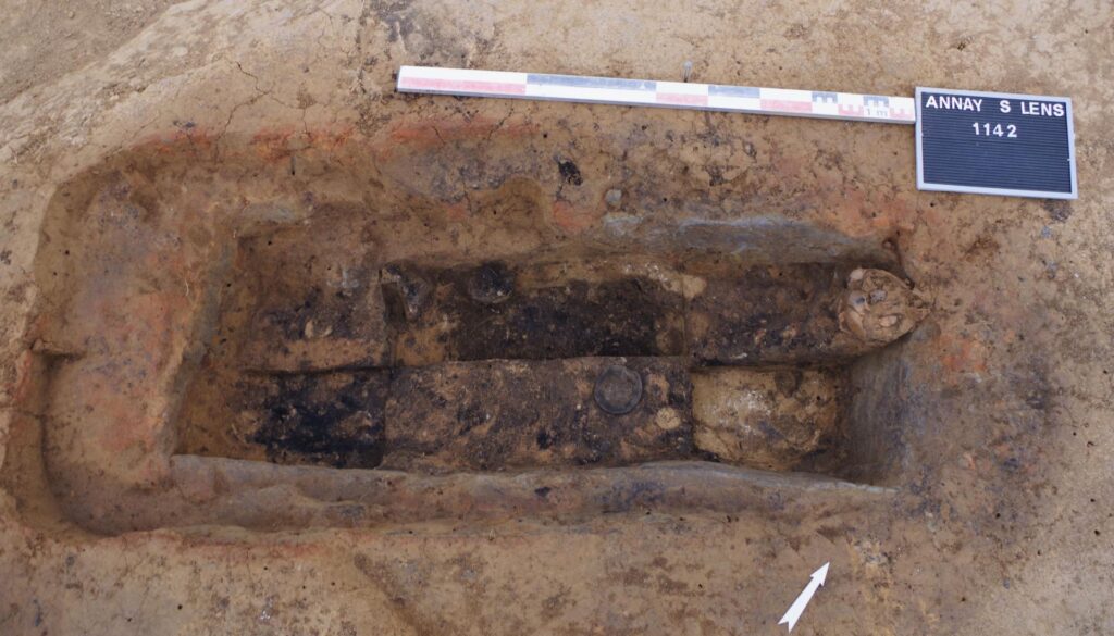 Άποψη από ψηλά ενός ορθογώνιου αρχαιολογικού λάκκου που περιέχει τμήματα ανθρώπινου σκελετού, κεραμικά αγγεία και ίχνη οργανικής ύλης. Δίπλα υπάρχει μια κλίμακα μέτρησης και μια πινακίδα με την ένδειξη "ANNAY S LENS 1142".