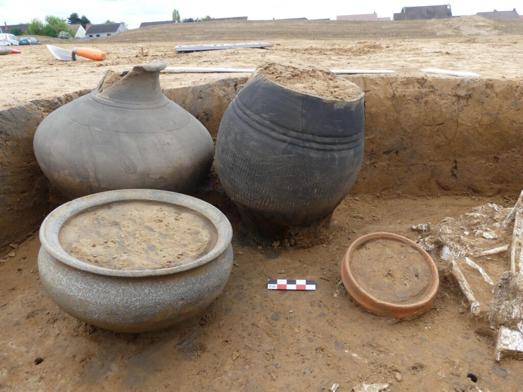 Τέσσερα μεγάλα πήλινα αγγεία (στάμνες και λεκάνες) σε γκρίζο και καφέ χρώμα, τοποθετημένα μέσα σε ένα σκάμμα. Στο βάθος διακρίνονται οικίες και ο εξοπλισμός των αρχαιολόγων.