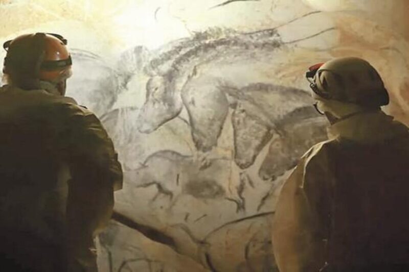 Δύο ερευνητές με προστατευτικό εξοπλισμό και φακούς κεφαλής στέκονται μέσα το σπήλαιο Σοβέ και παρατηρούν αρχαίες βραχογραφίες που απεικονίζουν μια ομάδα αλόγων σχεδιασμένων με κάρβουνο πάνω στα πετρώματα.