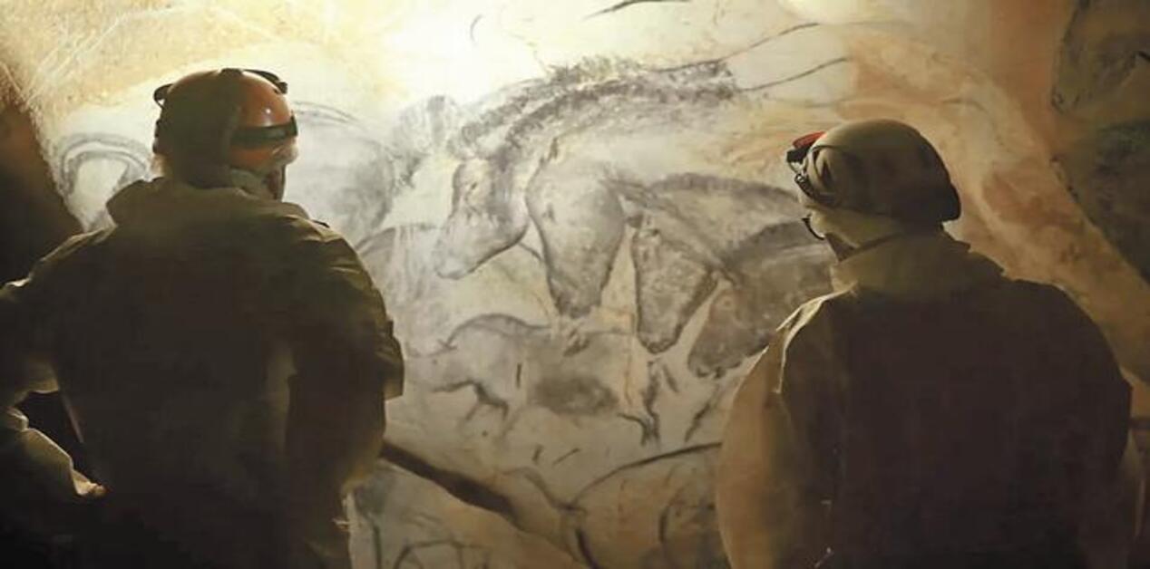 Δύο ερευνητές με προστατευτικό εξοπλισμό και φακούς κεφαλής στέκονται μέσα το σπήλαιο Σοβέ και παρατηρούν αρχαίες βραχογραφίες που απεικονίζουν μια ομάδα αλόγων σχεδιασμένων με κάρβουνο πάνω στα πετρώματα.