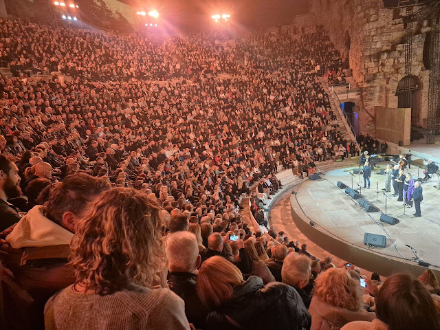 Άποψη από τις κερκίδες του Ηρωδείου κατά τη διάρκεια συναυλίας, με το κοινό σε πρώτο πλάνο και τη φωτισμένη σκηνή στο βάθος. Διακρίνονται οι καλλιτέχνες στη σκηνή και το ιστορικό πέτρινο τείχος υπό θερμό φωτισμό.