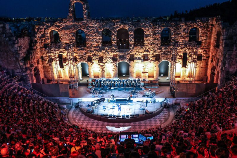 Πανοραμική νυχτερινή άποψη του Ωδείου Ηρώδου του Αττικού γεμάτου με κοινό κατά τη διάρκεια συναυλίας. Στη φωτισμένη σκηνή διακρίνονται μουσικοί και χορωδία, ενώ στο βάθος υψώνεται το επιβλητικό πέτρινο τείχος της αρχαίας σκηνής με τις τοξωτές εισόδους.