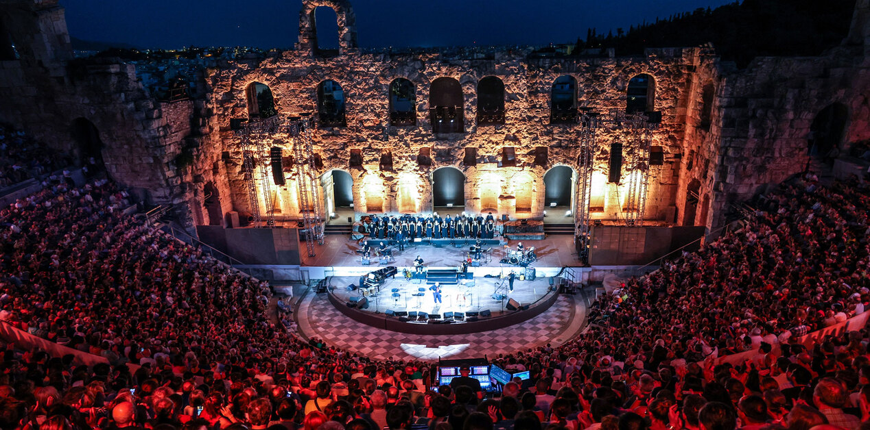 Πανοραμική νυχτερινή άποψη του Ωδείου Ηρώδου του Αττικού γεμάτου με κοινό κατά τη διάρκεια συναυλίας. Στη φωτισμένη σκηνή διακρίνονται μουσικοί και χορωδία, ενώ στο βάθος υψώνεται το επιβλητικό πέτρινο τείχος της αρχαίας σκηνής με τις τοξωτές εισόδους.