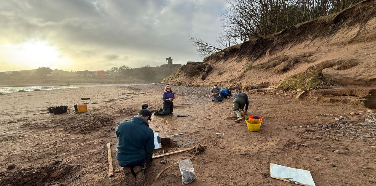 Μια ομάδα αρχαιολόγων και ερευνητών εργάζεται σε μια παραλία με λάσπη και άμμο κάτω από έναν συννεφιασμένο ουρανό. Οι ερευνητές είναι γονατισμένοι και ανασκάπτουν προσεκτικά το έδαφος χρησιμοποιώντας εργαλεία και κουβάδες, ενώ στο βάθος διακρίνεται ένας απόκρημνος χωμάτινος λόφος και ένα ιστορικό κτίσμα.
