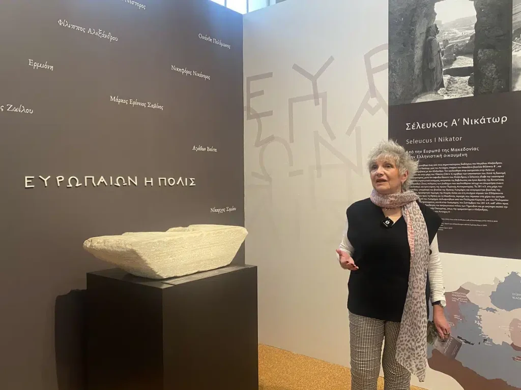 Η Δρ. Γεωργία Στρατούλη μιλάει δίπλα σε ένα πέτρινο αρχαιολογικό εύρημα στην έκθεση για την αρχαία Ευρωπό.