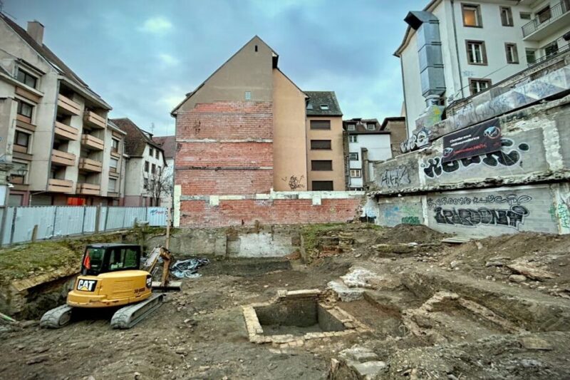 Μια ευρεία λήψη ενός αστικού αρχαιολογικού χώρου στο Bas-Rhin της Γαλλίας, με έναν κίτρινο εκσκαφέα σε ένα σκάμμα που περιβάλλεται από κτίρια με γκράφιτι.