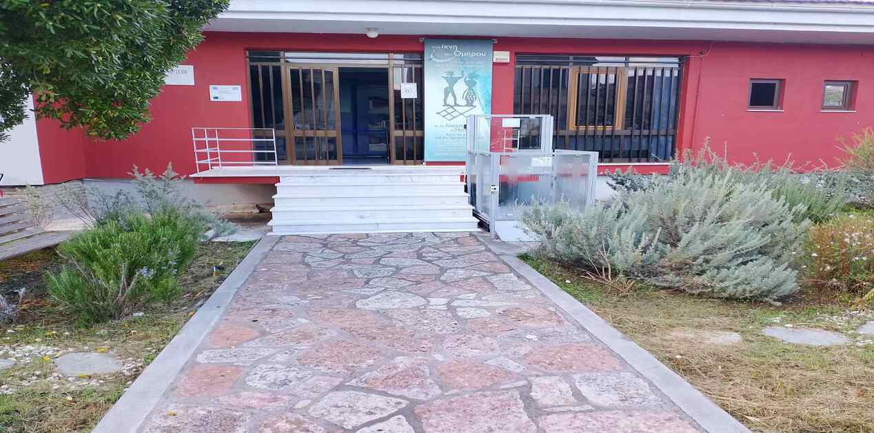 Εξωτερική άποψη της εισόδου του Αρχαιολογικού Μουσείου Βαθέος Ιθάκης με κόκκινους τοίχους, μαρμάρινα σκαλιά, πλακόστρωτο διάδρομο και ειδικό ανελκυστήρα ΑμεΑ.